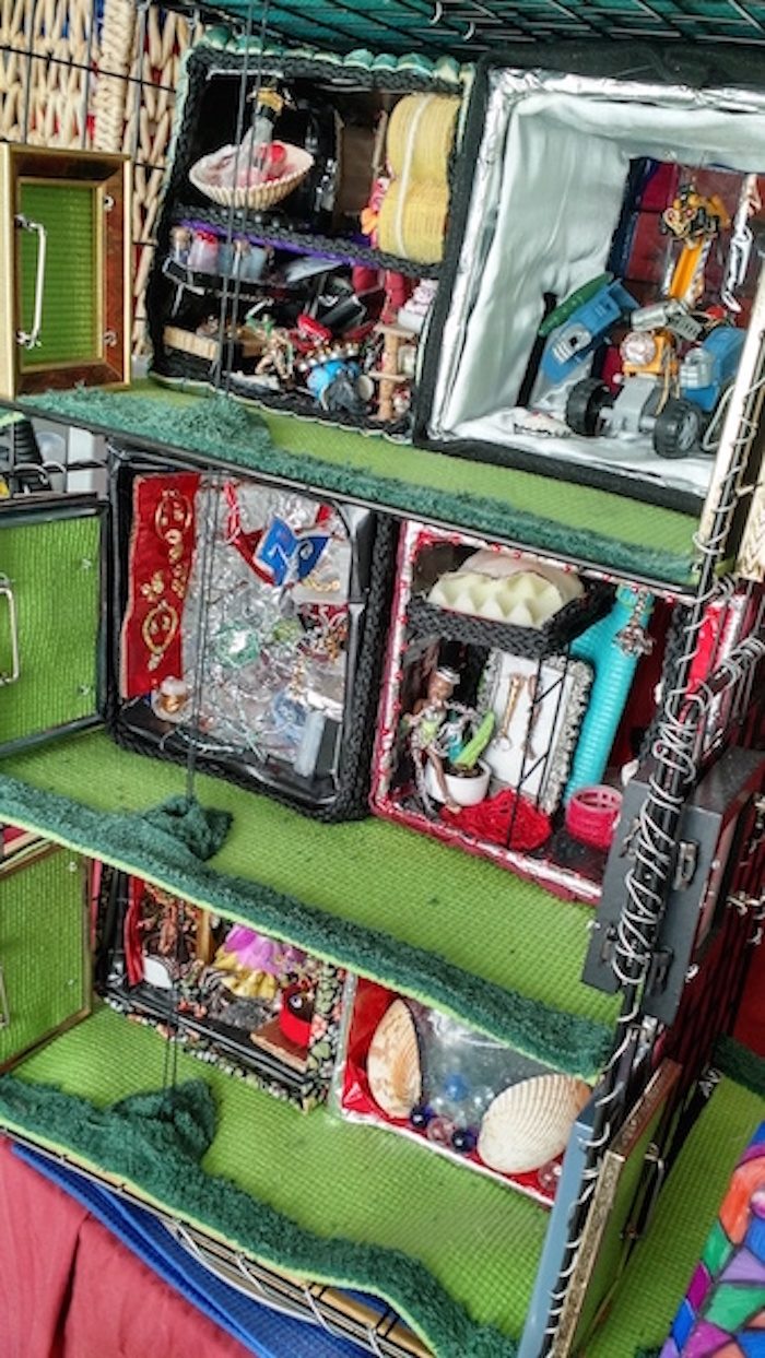 Another still photo of half-scale model of 6 apartment cubes on 3 stories of wire shelving. Can see that the top 2 apartments are a densely-furnished 2-story cube and a bare 1-room cube with a large robot in it. Second two are a mostly-silver room with a red display of tiny jewelry on one side, and a room with a humanoid doll standing under a raised bunk bed, but not clear enough to see much more. Some seashells barely visible in the right-side lowest unit.