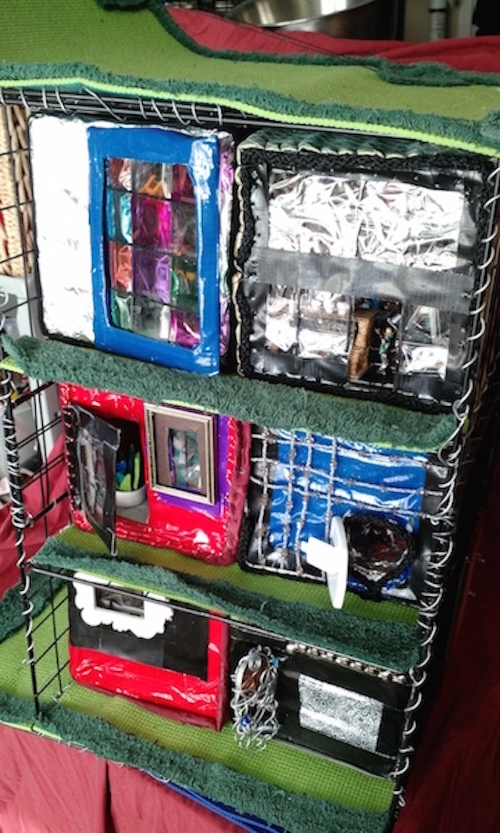 Another still photo of half-scale model of 6 apartment cubes on 3 stories of wire shelving. This one is focused on the outer side. Top two cubes show a large faux-stained-glass door and a clear wall with one small door. Second two are a red cube with a rectangular door and window, and a blue cube with one small hatch-like door and the rest decorated with strings of beads. Bottom two: a red cube with 1 window and no door, and a black-and-white one with an ornate wire door holding 1 colored circle of glass.