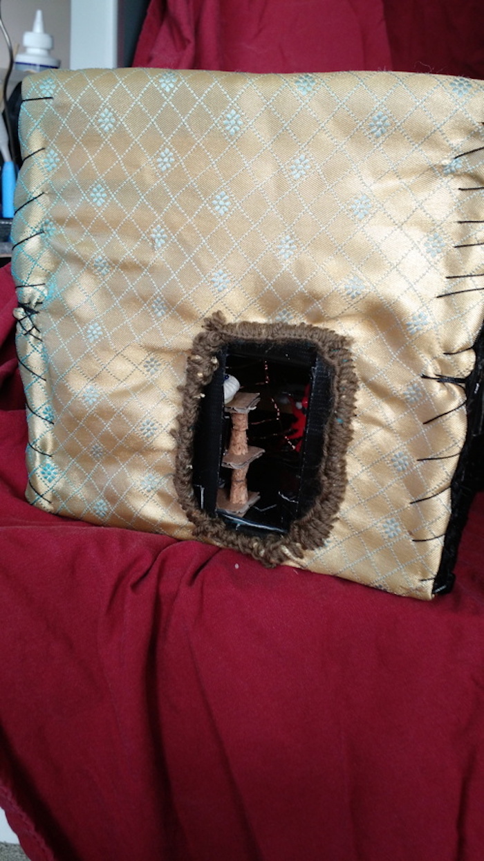 View of the Baking Cooperative mockup cube, removed from the shelving, seen from the side. Outer wall is clad in a golden, patterned fabric, with one open doorway hemmed with hand-sewn brown embroidery floss. This is the side door that leads into Operator 16's cube.