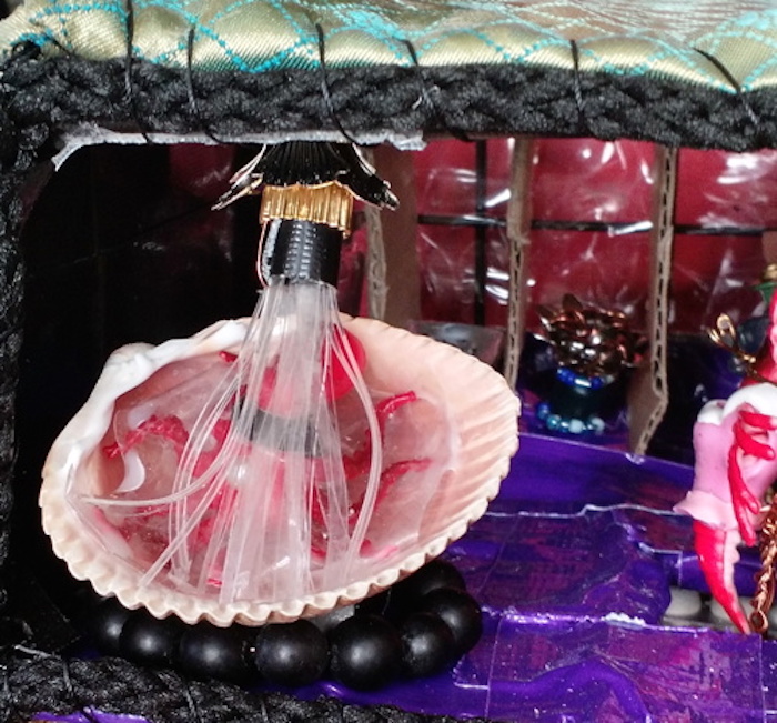 Another close-up of the upstairs bathroom in the Baking Cooperative mockup cube, this time focusing on the bathtub. In this version it is on the floor in front of the stalls, and it's made of a scallop shell on a ring of black beads. The shower stream from the nozzle above is made of thin strips of clear plastic, and a tiny octopus rests beneath the spray.