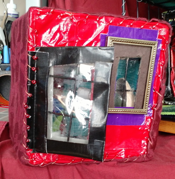 Photo of the sample mockup of Sessaris's apartment, removed from the shelving, seen from the closed side. This angle shows that the cube's outer back wall is shiny red, decorated with copper wire and red beads around the edges. A window is set in a small gold picture frame. A black-framed door, made mostly of clear windows, opens on a hinge also made of copper wire and red beads.