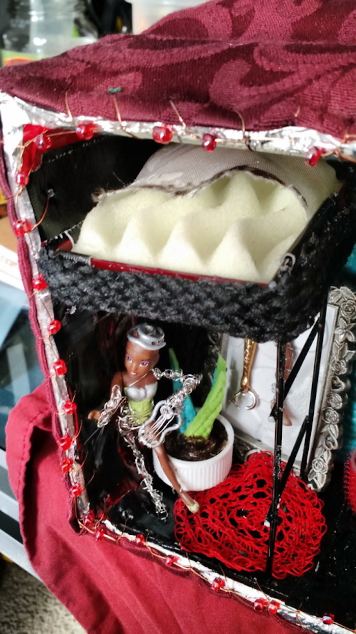 Photo of the sample mockup of Sessaris's apartment, removed from the shelving, seen from the open side. Another view of Sessaris, standing under the bunk bed, showing more of the bed from above. It is made of plain off-white mattress foam, and the sides of it are decorated with black rope. This angle shows that the cube's front is also decorated with copper wire and red beads around the edges.