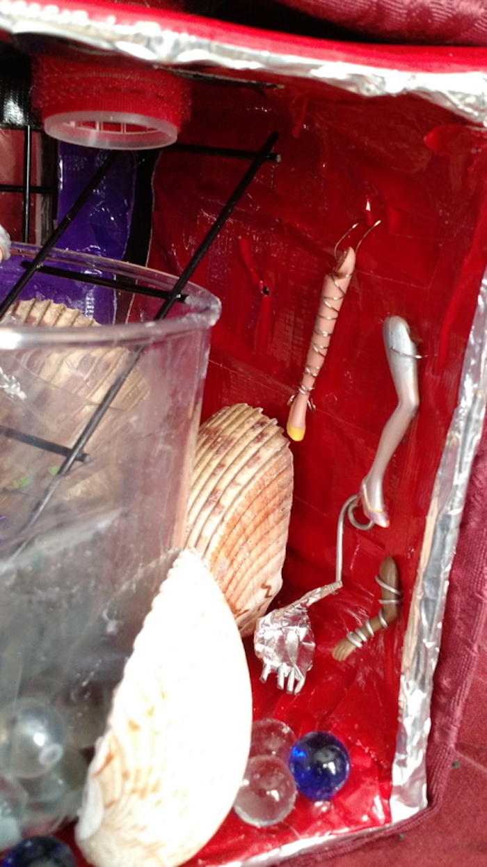 Photo of the sample mockup of Kit's apartment, removed from the shelving, seen from the open side. This view focuses on the right-side wall and the assortment of prosthetic limbs that hang there: a pink-skinned realistic leg wrapped in wire with a yellow shoe, a similar leg painted completely silver, a brown-skinned arm wrapped in wire, and a skinny metal leg with a large, bear-like clawed foot.