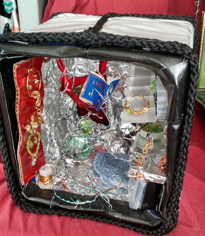 Photo of the sample mockup of the apartment, removed from the shelving, seen from the open side. The rough-textured walls, floor and ceiling in this version are made from aluminum foil. In this version the stand with jewelry is hanging just inside the cube, on the left wall. It is made of red satin and covered with brass jewelry set with green stones. The workbench is attached to the right wall, and rests on cabinets full of various marbles and beads, made of small translucent square snap-top containers. Above it, the row of cubbies for jewelry-making supplies are made of a 7-day pill container, with all the snap tops open to form a long shelf in front of the cubbies. The creature at the workbench is made of a large roundish piece of green glass vase-filler, and its tentacles are made of thick steel wire with thinner steel wire wrapping around it, holding small green crystal beads against it in places. One of the two other similar creatures is is leaving the apartment, the other is resting in a red satin hammock reading a small blue book made of stapled paper.