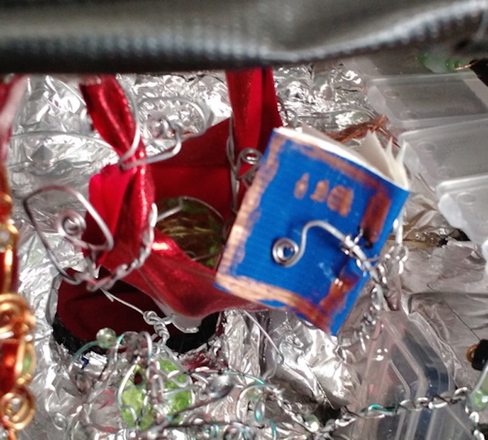 A close-up of the creature resting in the hammock. Its book has a cover made of a piece of blue duct tape, with marks in gold ink that do not spell anything in any recognizable language. It is holding the book in two branches of one of its steel tentacles. The other tentacles wrap around the ends of the hammock, holding onto them like the ropes of a swing. Its body, this one made from a large green faceted bead, is almost hidden in the depths of the hammock.