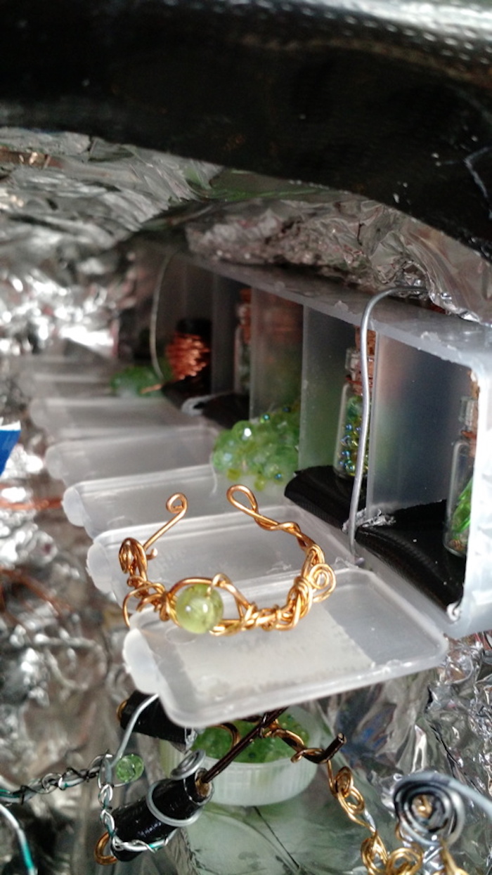 A close-up of the row of cubbies for jewelry-making supplies, attached to the wall above the workbench. They are made of a 7-day pill container, with all the snap tops open to form a long shelf in front of the cubbies. On that shelf rests one piece of brass jewelry, perhaps a headband, with a round green bead on it. The mini-bottles in the cubbies hold tiny green beads. One of the cubbies also contains a pile of loose green beads, and another contains a roll of copper wire. 