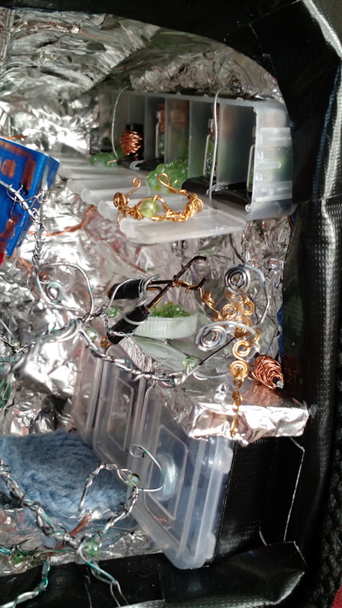 A close-up of the creature making jewelry at the workbench. In one of its tentacles, it holds a pair of pliers made from brass wire, with hand-grips made from black tape. In the other it holds a length of brass chain it has just made, with each link an S-shape of spiraling wire. Another roll of copper wire and a bottle-cap full of green crystal beads are sitting on the workbench.