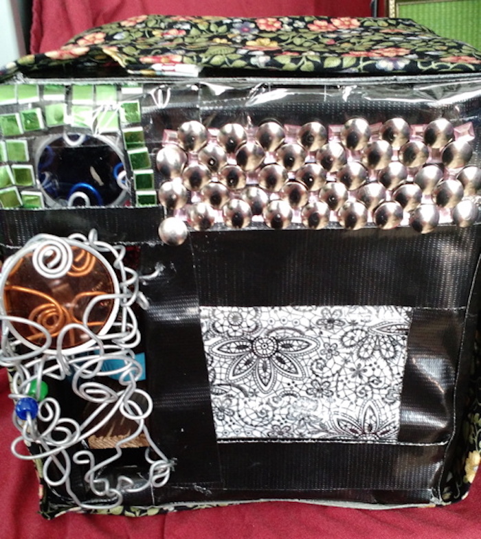 Photo of the sample mockup of the apartment, removed from the shelving, seen from the back. This view shows the back door, which in this version is made of randomly twisted steel wire with a few beads and a round orange camera filter incorporated into it for a window. Above it, a similar blue camera filter forms another window, surrounded by green stick-on tiles, while the remainder of the upper wall to the right of that is tiled with round silver thumbtacks. Below that, the rest of the wall is covered in black tape and black-and-white floral-patterned tape.