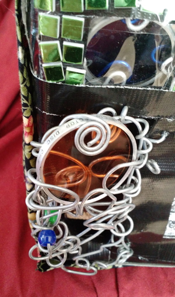 Photo of the sample mockup of the apartment, removed from the shelving, seen from the back. This view focuses more closely on the back door, made of randomly twisted steel wire with a few beads and a round orange camera filter incorporated into it for a window. A large spiral of wire at the top holds the camera filter in place. It is made of orange glass in a circular aluminum frame, and in this view you can see some words engraved on the frame, including CORRECTOR DISC.