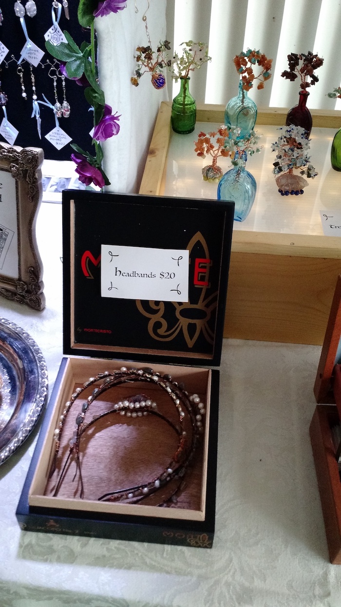 close-up of a box of jeweled headbands. In the background is a platform covered with small decorative trees and flowers, made from wire and gemstone chip beads, some with roots wrapped around stone bases, and some arranged in miniature glass bottles.